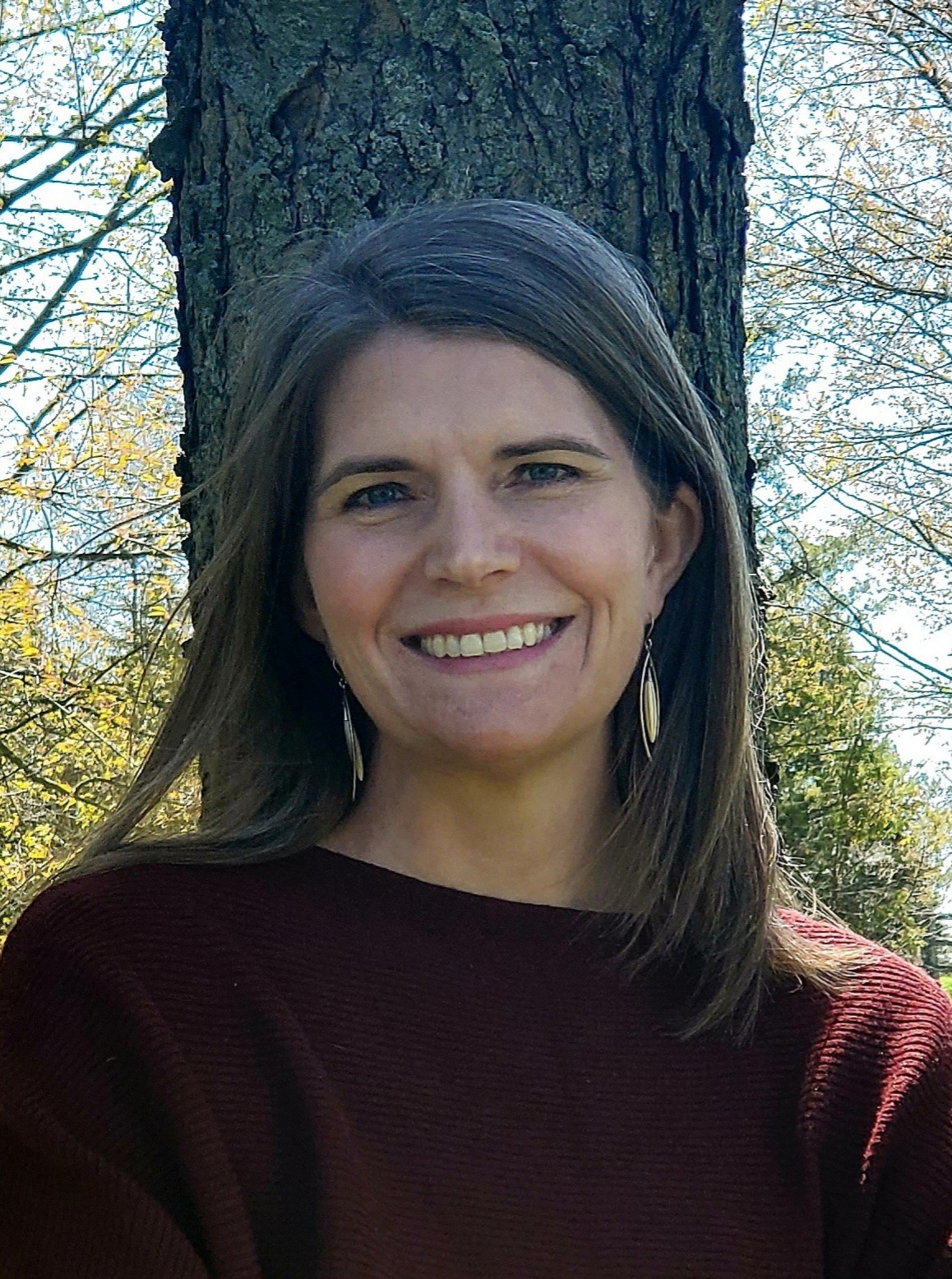 Picture of a woman with long brown hair and a friendly smile standing in front of a tree outdoors